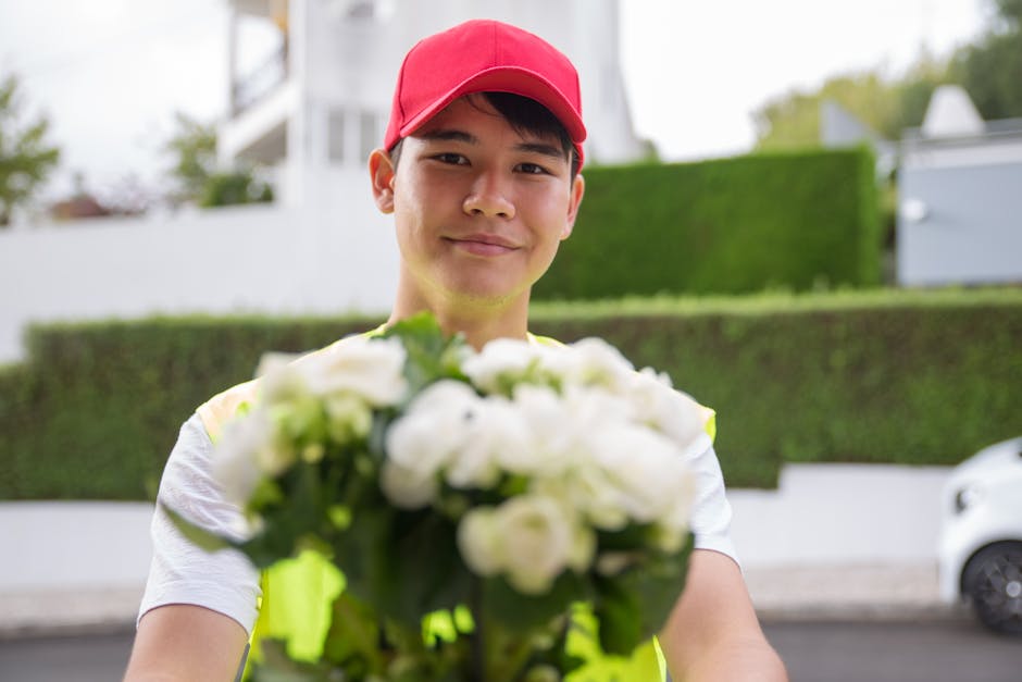 A young male flower delivery courier wearing a red cap and a light-colored T-shirt, smiling as he holds a vibrant bouquet of white roses and greenery towards the camera. The bouquet is wrapped with a
