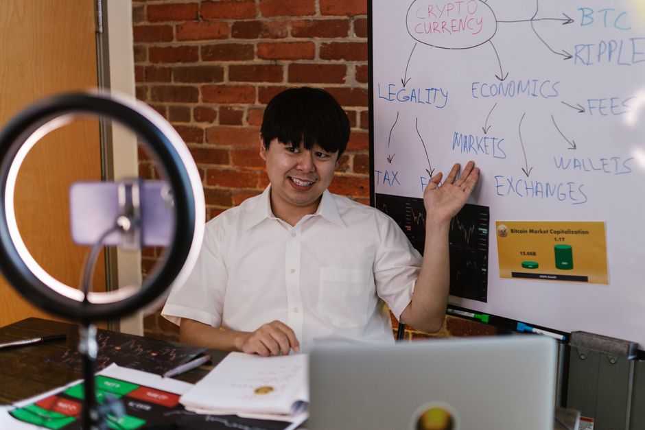 A young man with black hair wearing a white shirt sits at a desk in an office setting, demonstrating a floral arrangement or bouquet. He is smiling and gesturing with his right hand towards a whiteboa