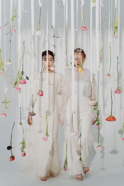 Two women stand behind a sheer, white curtain adorned with hanging pink, peach, and purple flowers with green stems, creating a delicate floral display. Both women are dressed in elegant, cream-colore