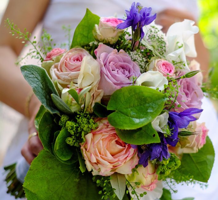 A vibrant floral bouquet featuring soft pink roses, white lisianthus, and striking purple irises, arranged with lush green leaves and delicate sprigs of baby's breath. The flowers are tightly clustere
