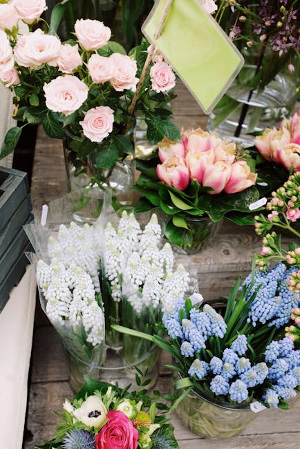 A variety of fresh floral arrangements displayed on a rustic wooden surface, featuring light pink roses with lush dark green foliage in a transparent glass vase, a bouquet of pink and white tulips wit