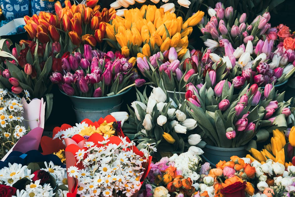 A vibrant display of fresh flower arrangements featuring a variety of tulips in colors including yellow, white, pink, purple, and orange, arranged in blue metal buckets. The tulips have smooth, slight